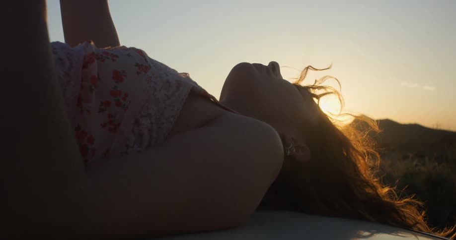 A woman is laying on a parked car during sunset