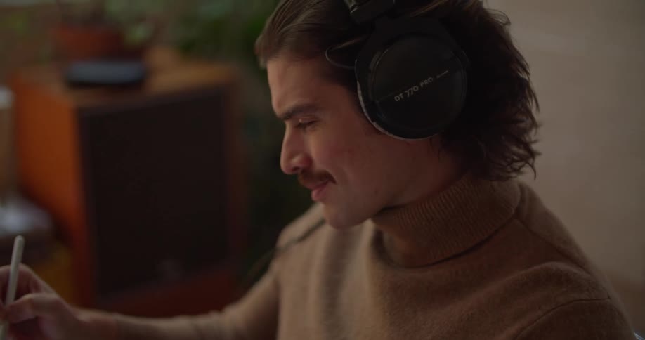 A man sitting down wearing headphones is holding a pen and writing as a record album spins on a turntable