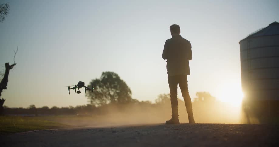 Silhouette of a man standing outside landing a drone and then picking it up and walking away as the sun shines brightly in the distance