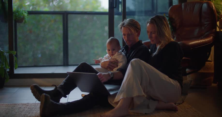 A woman is sitting on the floor typing on a laptop computer as her husband sits next to her holding their baby