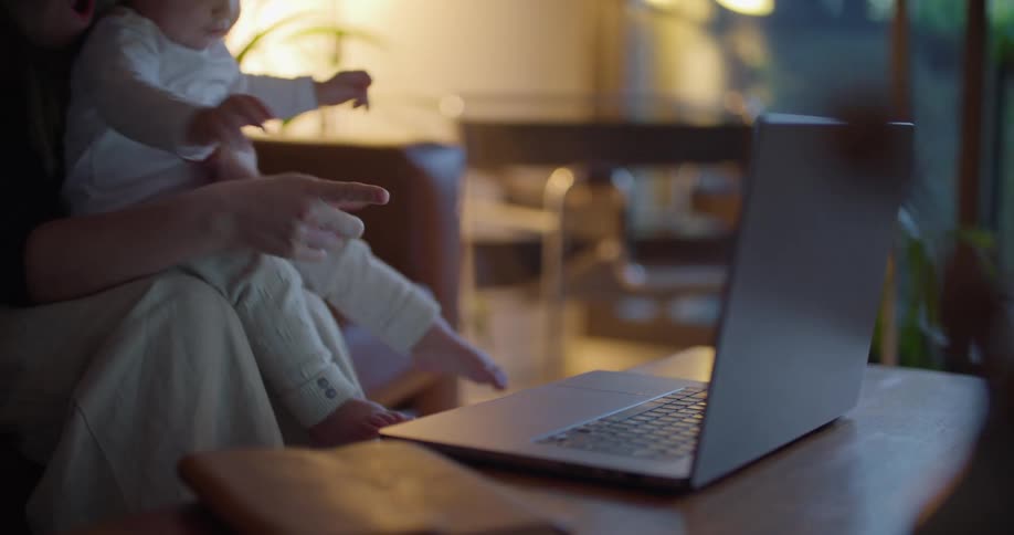 A woman is sitting on the couch holding her baby and showing her baby the laptop computer