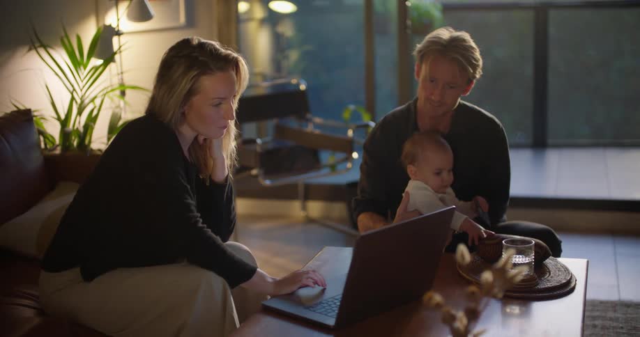 A woman is sitting on the couch looking at a laptop computer and showing her husband something as he sits on the floor holding their baby