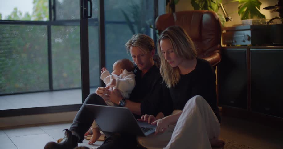 A woman is sitting on the floor typing on a laptop computer as her husband sits next to her holding their baby