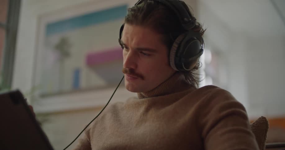 A man sitting down in front of a laptop computer and wearing headphones is holding a pen and thinking