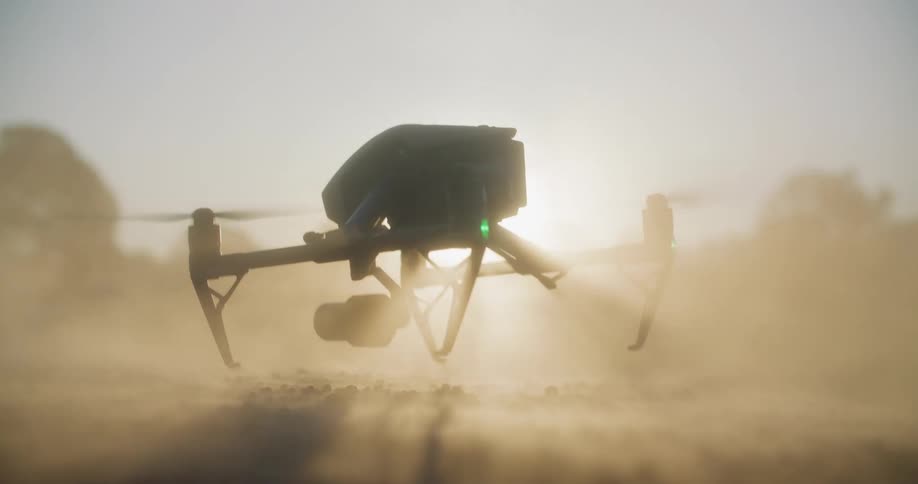 A drone is sitting on the ground as its propellers spin and dust flies as it takes off