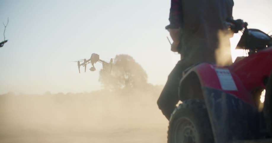 A man is operating a drone that is sitting on the ground as its propellers spin and dust flies as it takes off