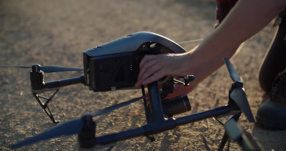 A drone is sitting on the ground and a man is attaching a camera to it