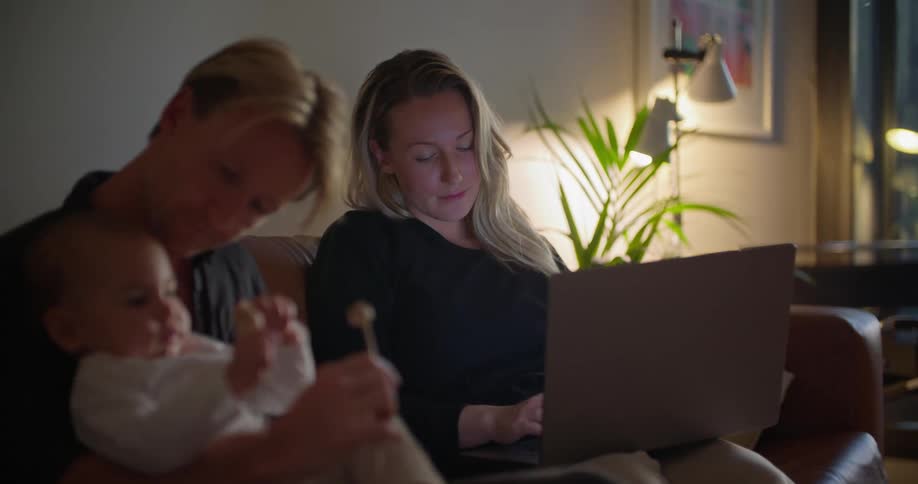 A woman is sitting on the couch typing on a laptop computer as her husband sits next to her holding their baby