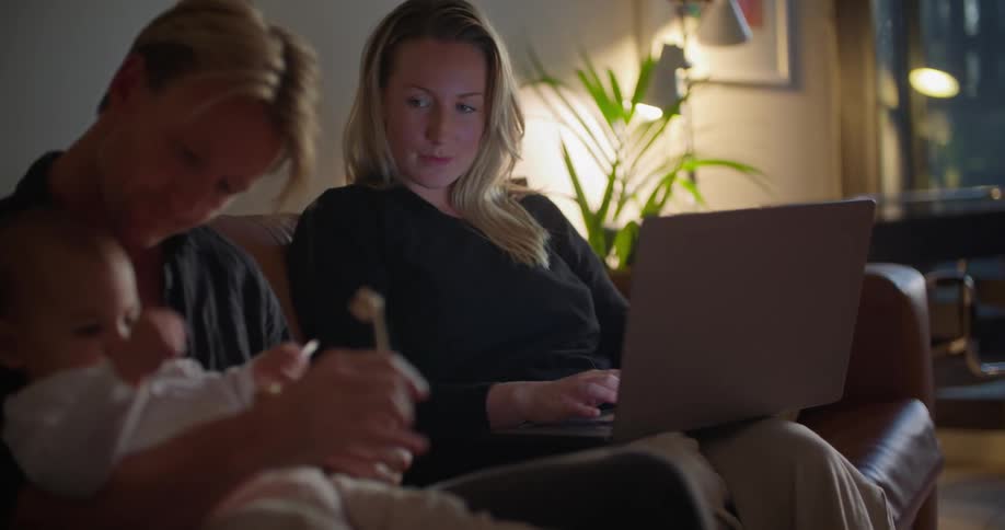 A man holds his baby on his lap as he sits next to his wife who uses her laptop and they talk and laugh