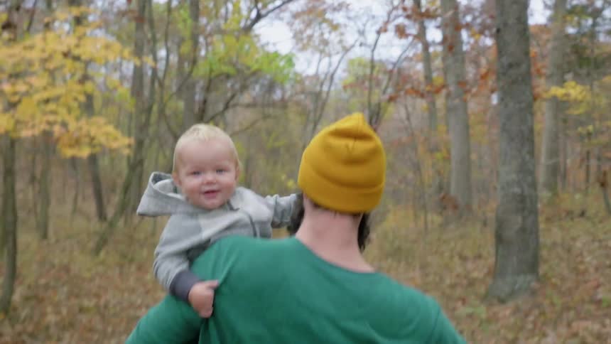 A man in a yellow hat is carrying a baby through a forest