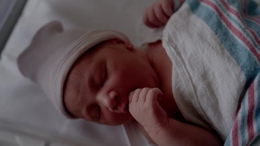A newborn baby suckles its hand as it lays in a hospital bassinet