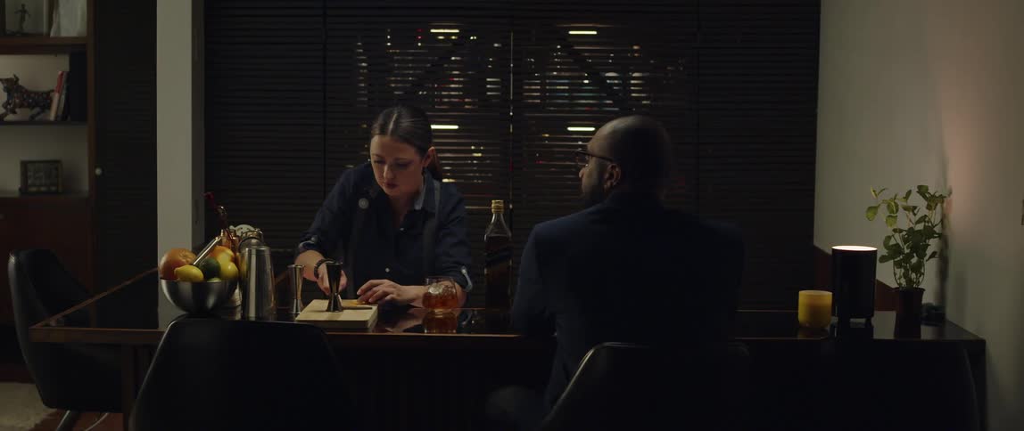 A bartender prepares the fruit for a drink then hands the drink to a customer sitting at the bar