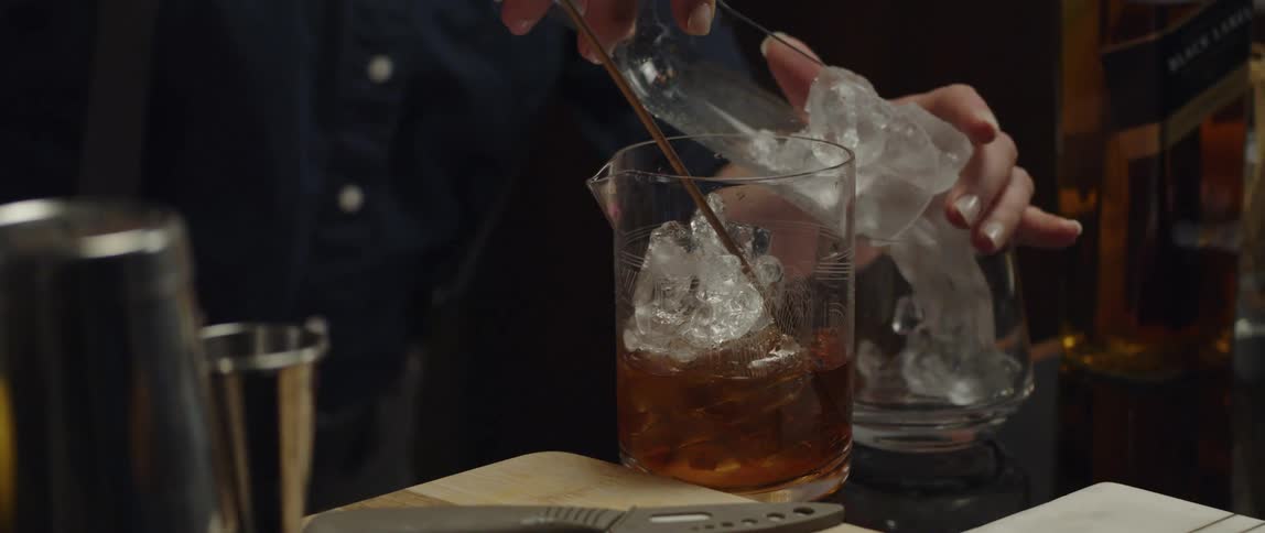 Female bartender preparing a drink pouring ice from a ice scoop into a glass at the bar counter
