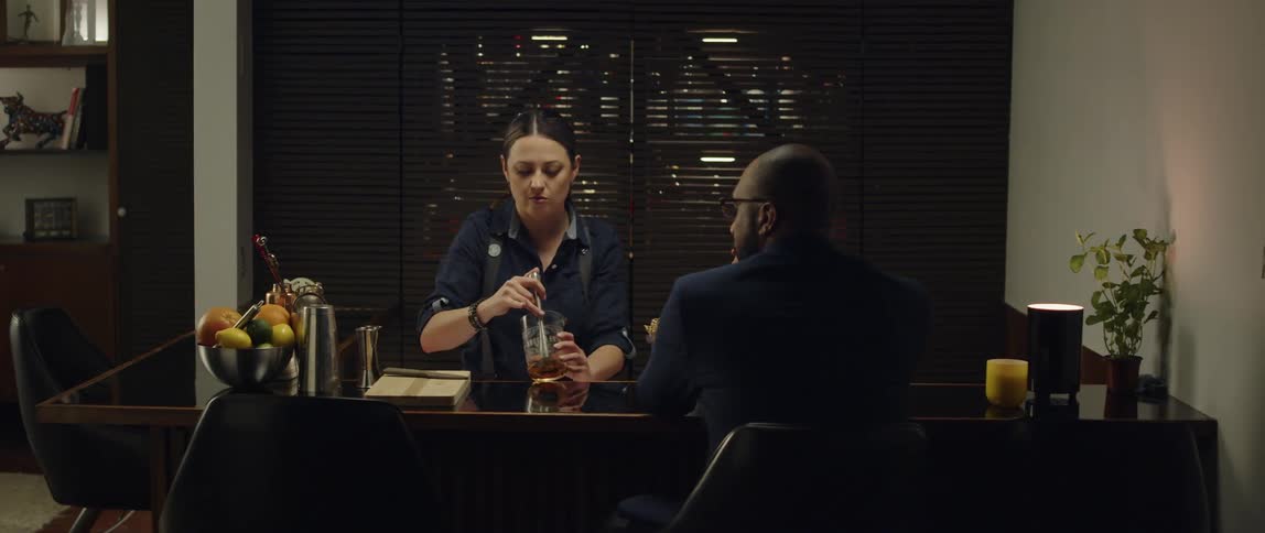 A man sits at a counter in a home as a woman mixes him a drink