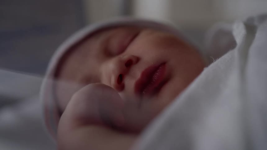 Baby with white blanket and hat lies in bed sleeping