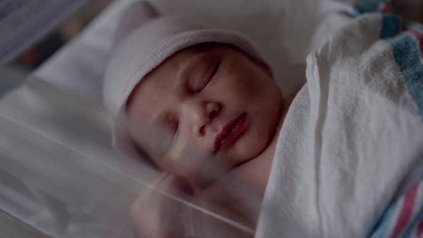 Newborn baby wearing a white cap sleeping on a plastic cradle under blankets