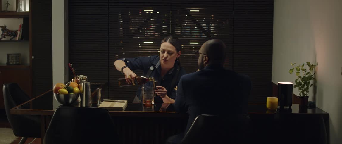 A man sits at a bar counter as a woman mixes a drink for him