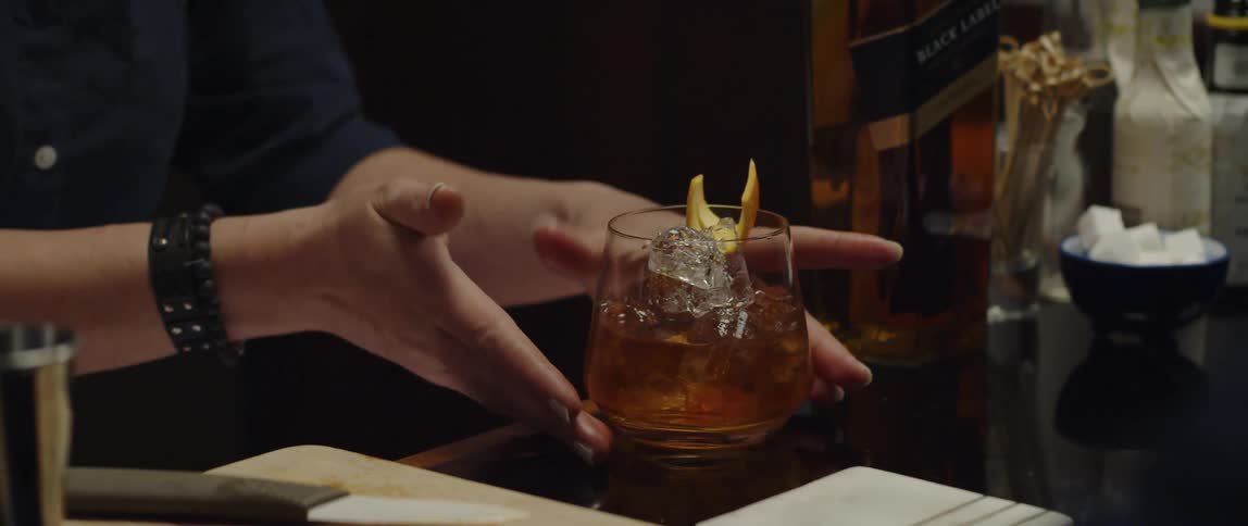 A bartender places an orange rind garnish on a glass then serves the drink at the bar on a napkin