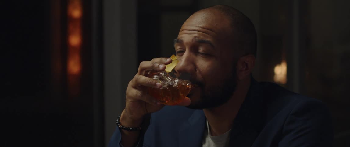 Man wearing a blazer drinking a glass of whiskey while talking to someone in a bar at night