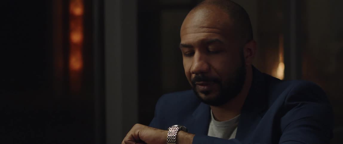 Man wearing blazer checks his wristwatch while sitting with his back to the fireplace