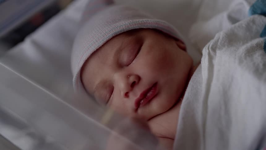Baby with pink hat and white blanket sleeps in bed