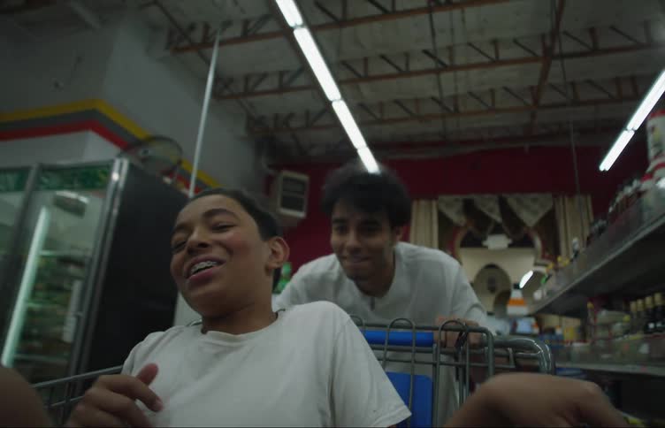 A teenage boy sitting inside a grocery cart throws food at the boy pushing the cart at a run