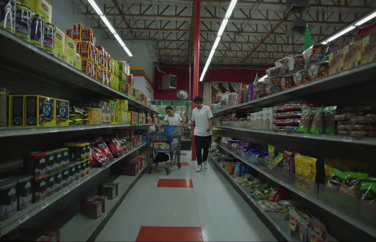 Two boys are pushing a shopping cart down the aisle of a grocery store and picking out snacks