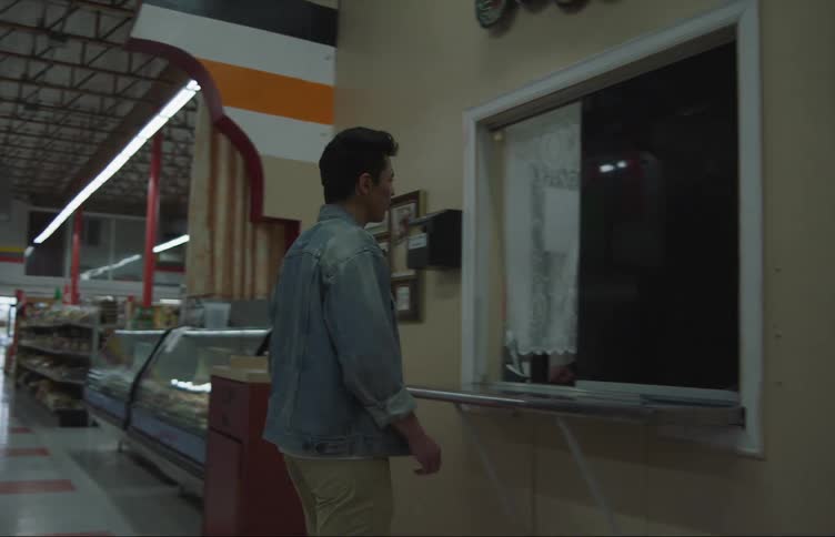 A man walks down a grocery store lane and briefly speaks to someone at a window covered with a curtain