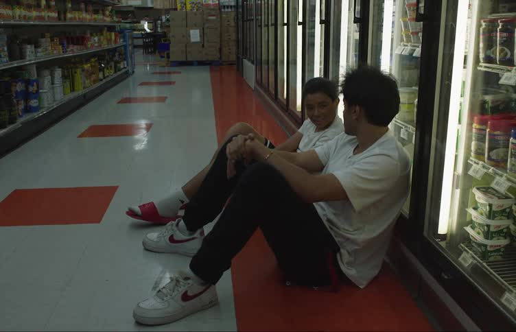 Two friends sit on floor in grocery store near refrigerated section