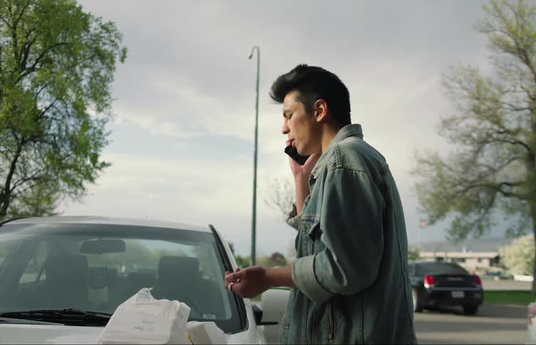 A man is standing outside by his car and eating food while talking on the phone