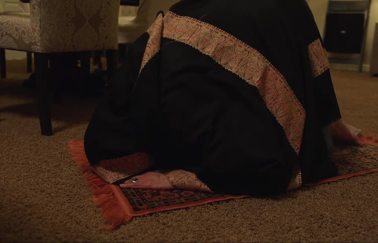 Muslim woman with black prayer dress performs prayer next to dining table in living room