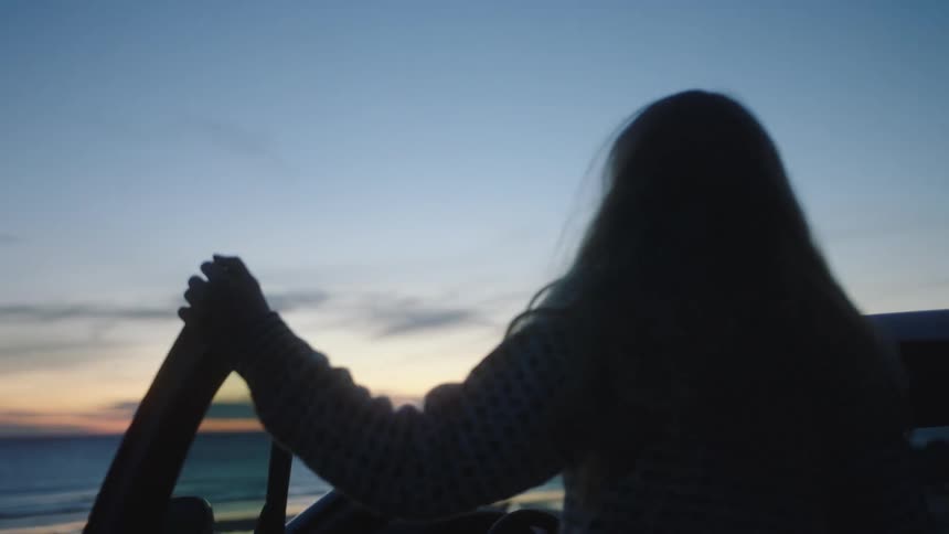 Woman gets out of car at beach as ocean stretches to the red tinted horizon