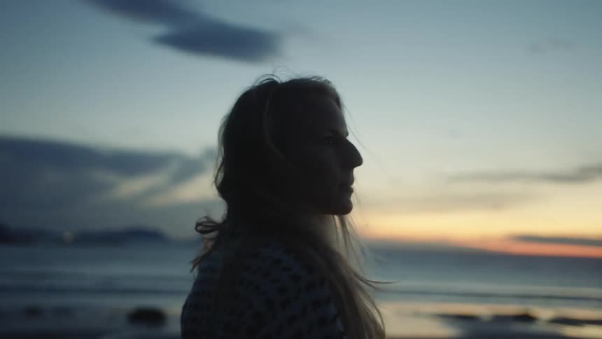 Blonde woman with long wavy hair is silhouetted against the sky at sunset on the beach