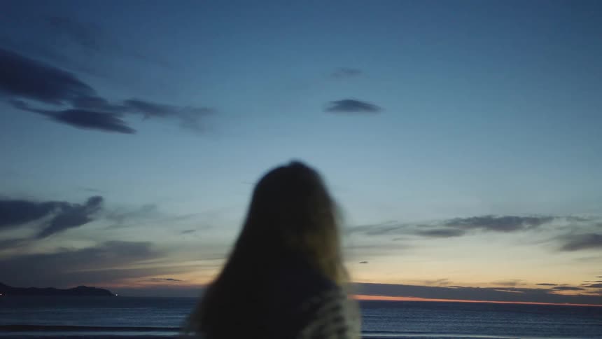 A woman standing on a beach slowly turns to face the ocean at sunset