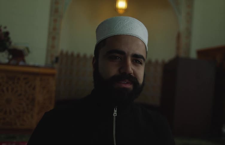 Imam with white skull cap beard and black jacket smiles at the mosque