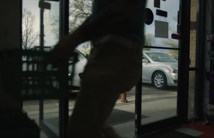 A man is sitting on a car hood eating as he talks on the phone and a worker moves product then a customer leaves the store with a bag