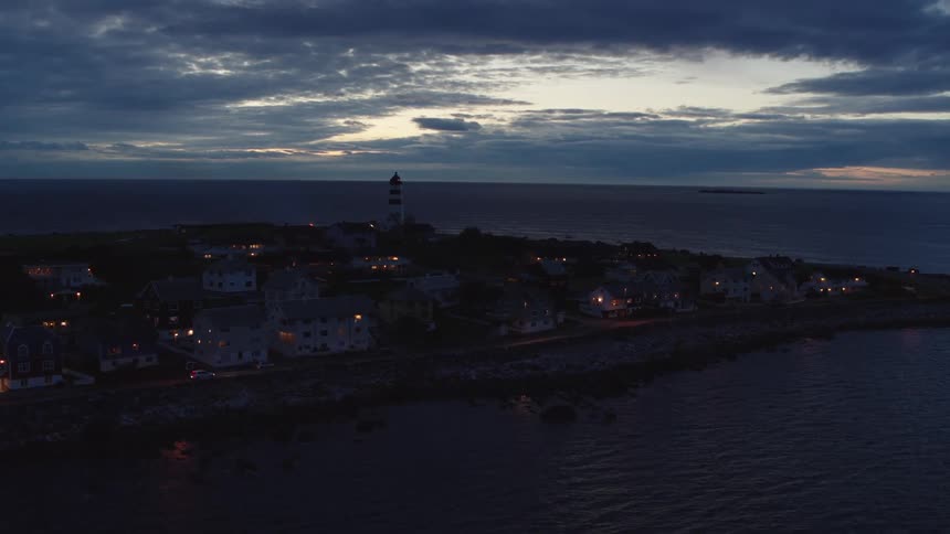 A lighthouse looms over a small town on a peninsula under a cloudy evening sky