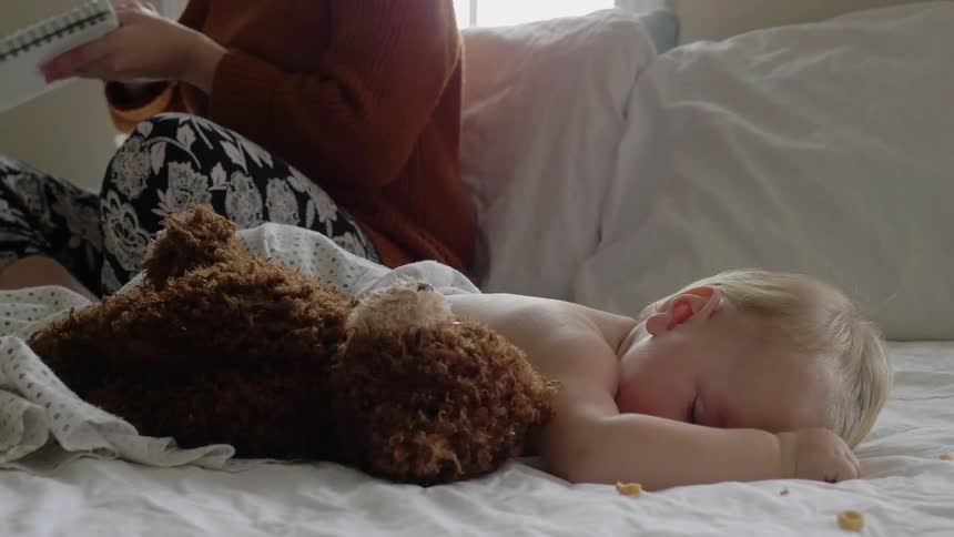 A woman sits on a bed as a baby sleeps next to a teddy bear