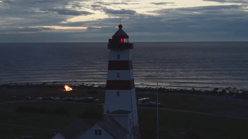 A lighthouse beacon glows red standing above a beach bonfire