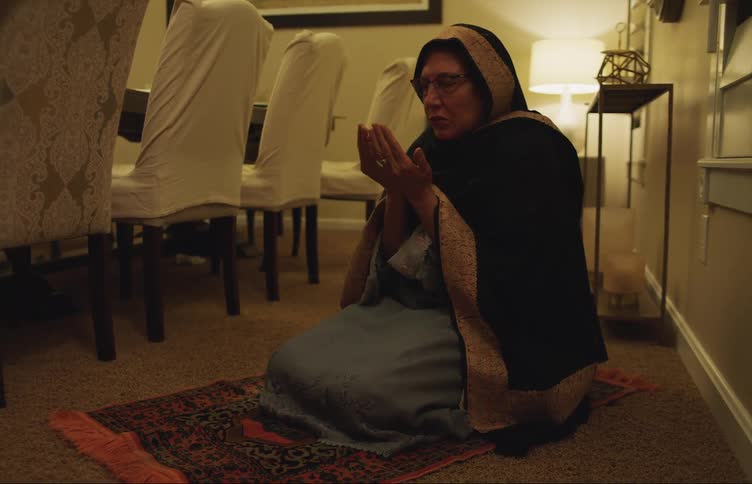 Middle aged muslim woman wearing a traditional veil praying while kneeling on a prayer rug in the dinner room at night
