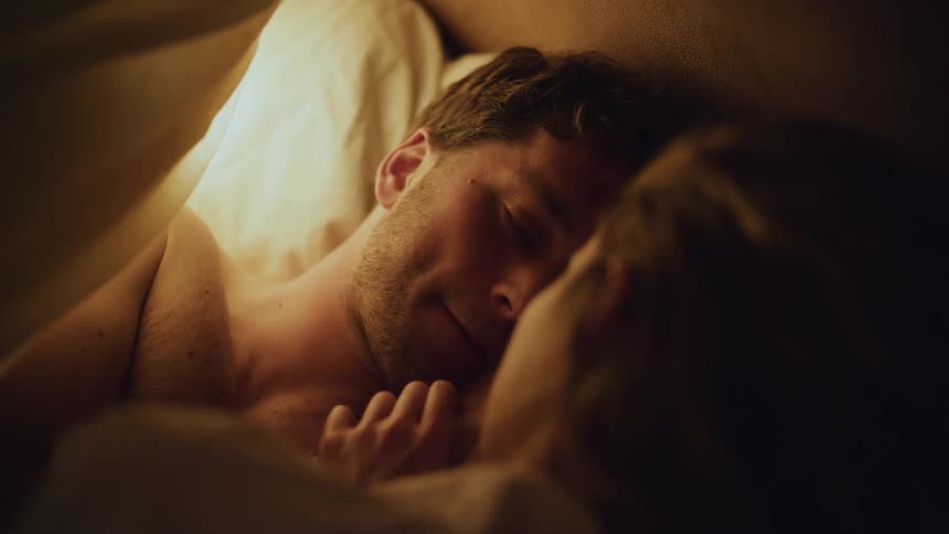 A man and woman lie in bed together and he looks at her and smiles and she rubs his chin