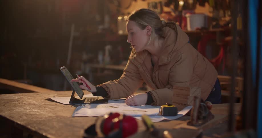 A woman leans on a table as she looks at a tablet then writes on some paper