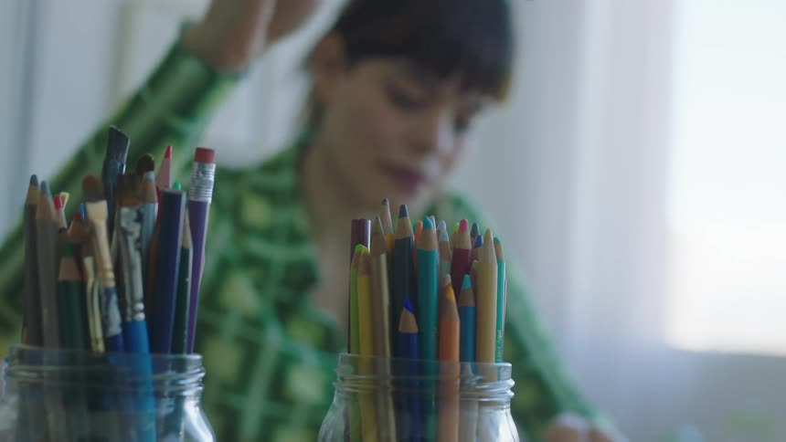 Two jars of colored pencils sit in front of a woman at a table and she chooses one to use