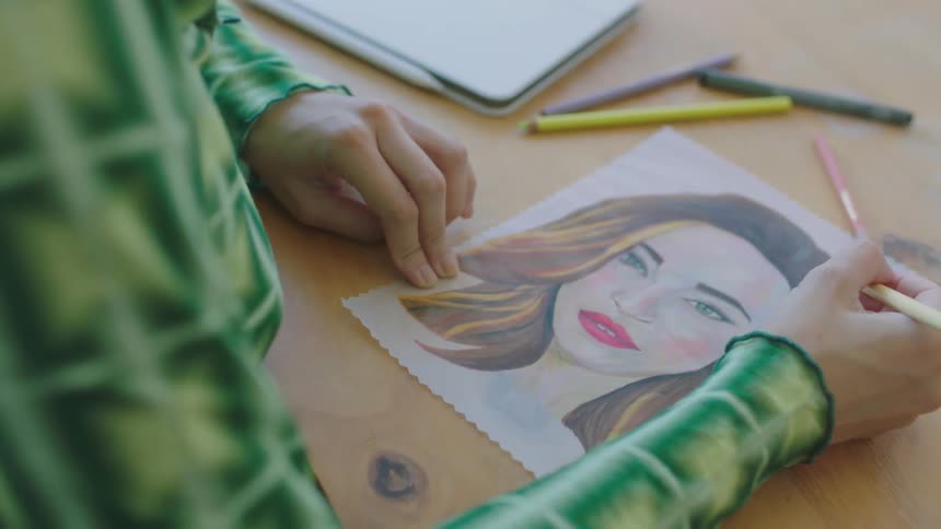 A woman colors a portrait using colored pencils while sitting at a desk indoors
