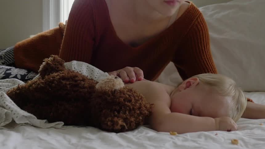 A baby is sleeping as a woman puts a teddy bear down