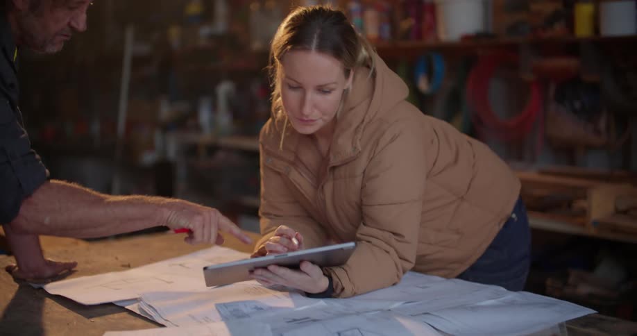 A man looks at a tablet a woman is holding as they stand over blueprints in a workshop