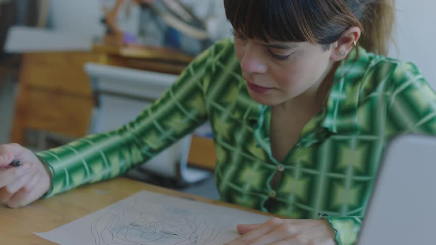 A woman uses a pen to draw on a sheet of paper while sitting at a table indoors