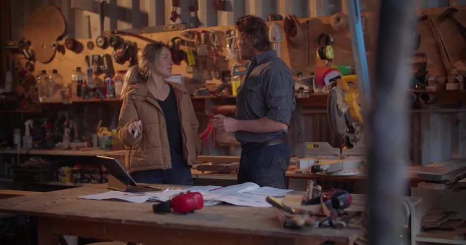 A woman and man talk with one another while referencing a computer tablet on its stand on a table in a workshop