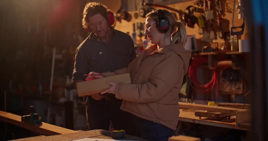 A man helps a woman with a building project inside of a workshop with a plethora of wood