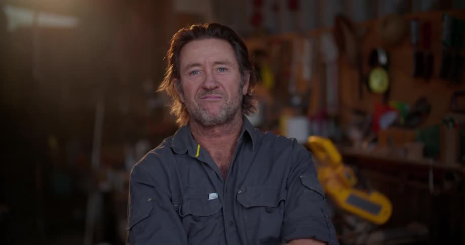 A man standing in a workshop smiles as he takes his glasses off